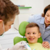 A dentist is teaching a small boy who is sitting on the dental chair, how to brush the teeth with brush and paste in his hands, a woman dentist can be seen around.