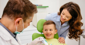 A dentist is teaching a small boy who is sitting on the dental chair, how to brush the teeth with brush and paste in his hands, a woman dentist can be seen around.