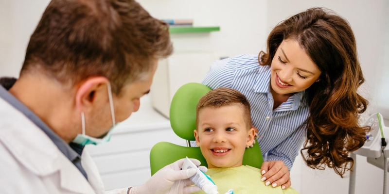 A dentist is teaching a small boy who is sitting on the dental chair, how to brush the teeth with brush and paste in his hands, a woman dentist can be seen around.