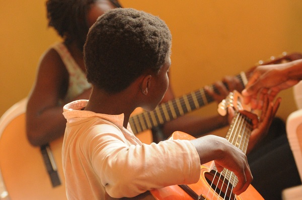 A child holds a guitar and learns how to play it from his teacher.