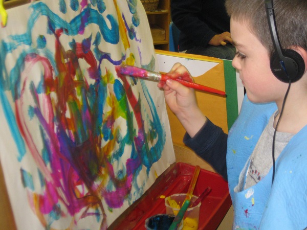 A small boy is painting on the drawing board with a painting brush on his hand, some paint is kept in the cup which can be seen straight below.